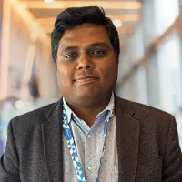 Portrait of a man in a dark blazer with a lanyard, standing in a bright hallway.